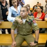 A man in military uniform sits on a wooden bench in a courtroom and is comforted by a woman.