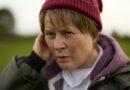 Monica Dolan, as Catherine Corless, touches her ear with a concerned expression outdoors