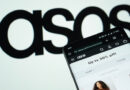 Photo of a smartphone open to the Asos website. The retailer's logo is displayed in black on a slightly off-focus white backdrop behind the phone.
